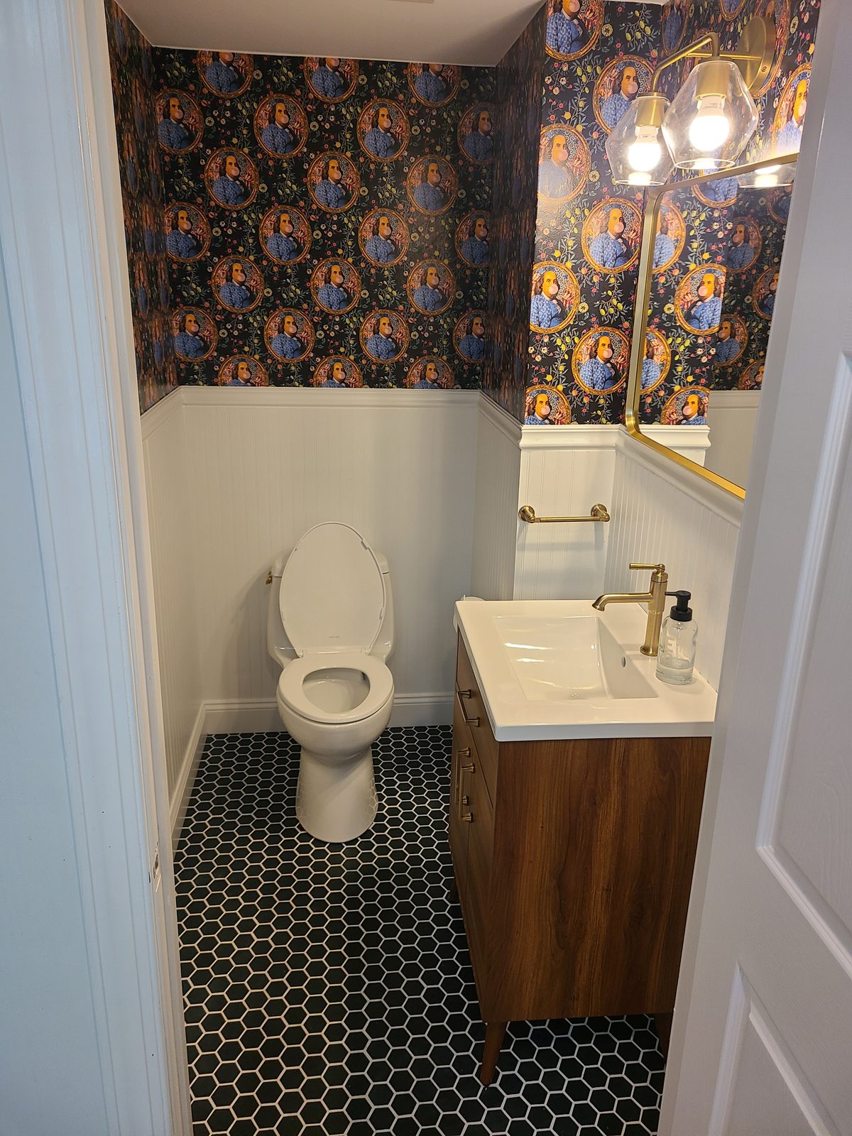 Remodeled powder room with bold portrait wallpaper, white beadboard wainscoting, black hexagon tile floor and a walnut vanity