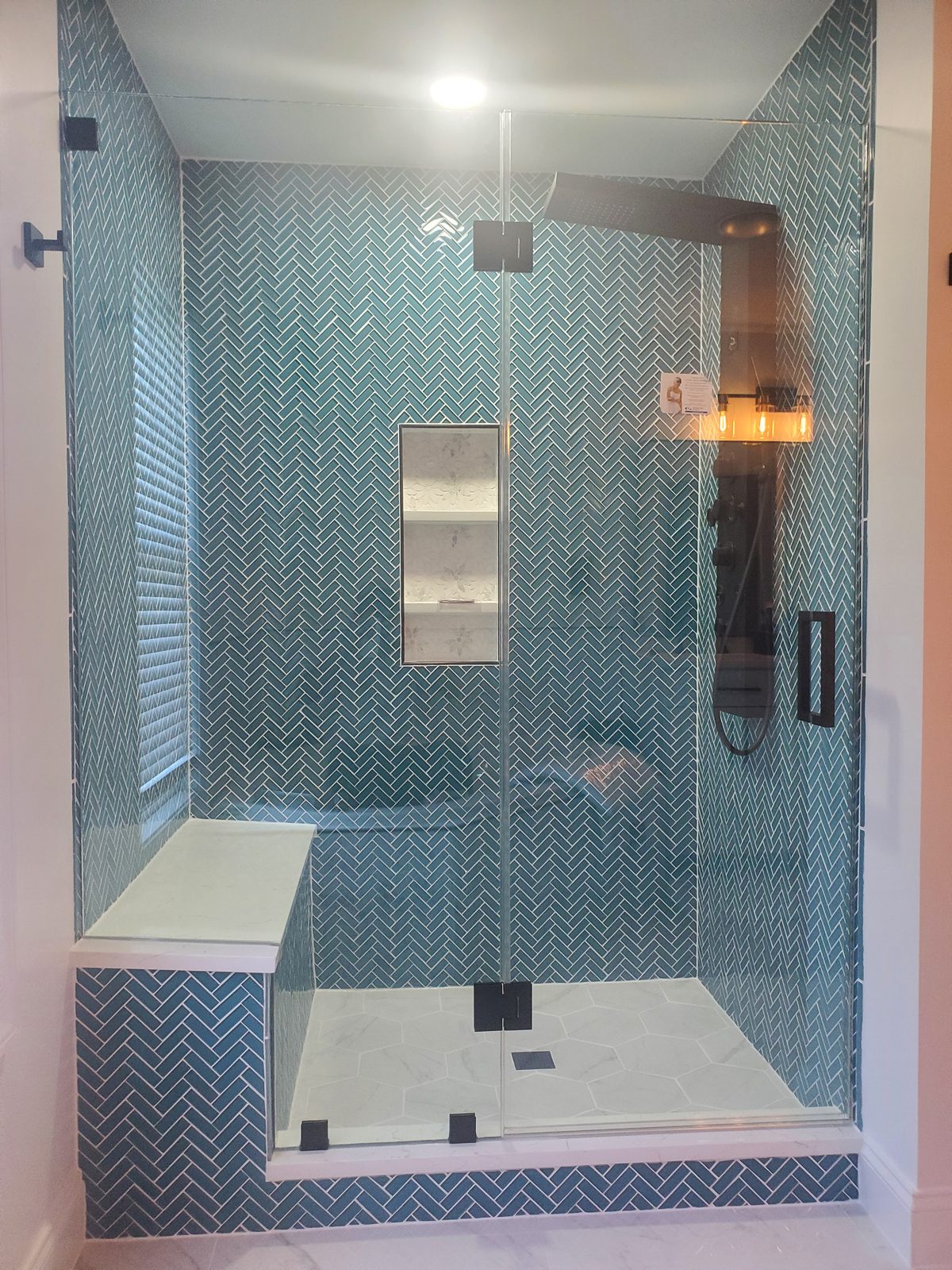 Rebuilt walk-in shower with teal herringbone glass tile, marble bench and niche, and a frameless glass door