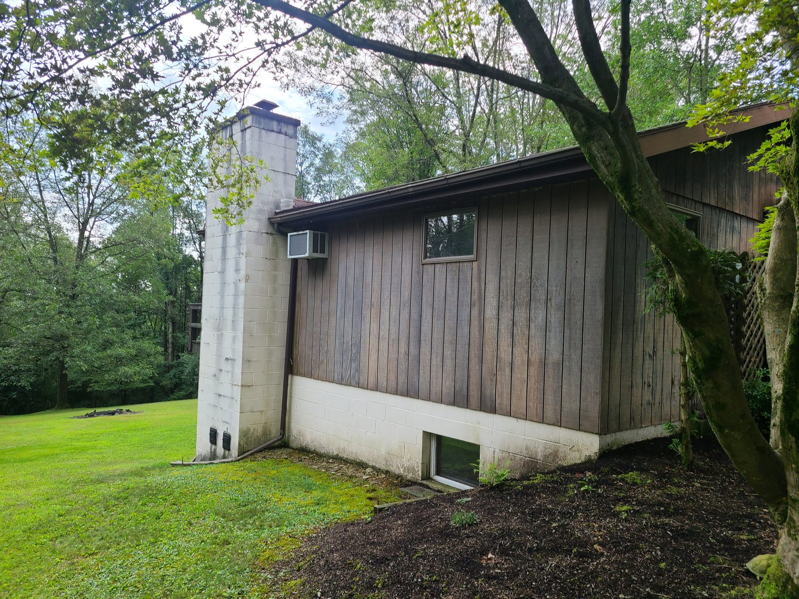 Side of the house before residing, with weathered vertical board siding, a block chimney and a window air conditioner